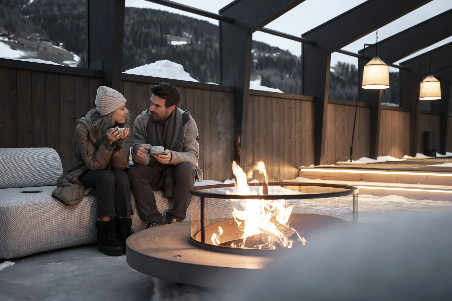 Hotel Monika in Sexten: Wohlbefinden inmitten der Dolomiten Paar sitzt draußen am Feuer und trinkt heiße Getränke im Winter