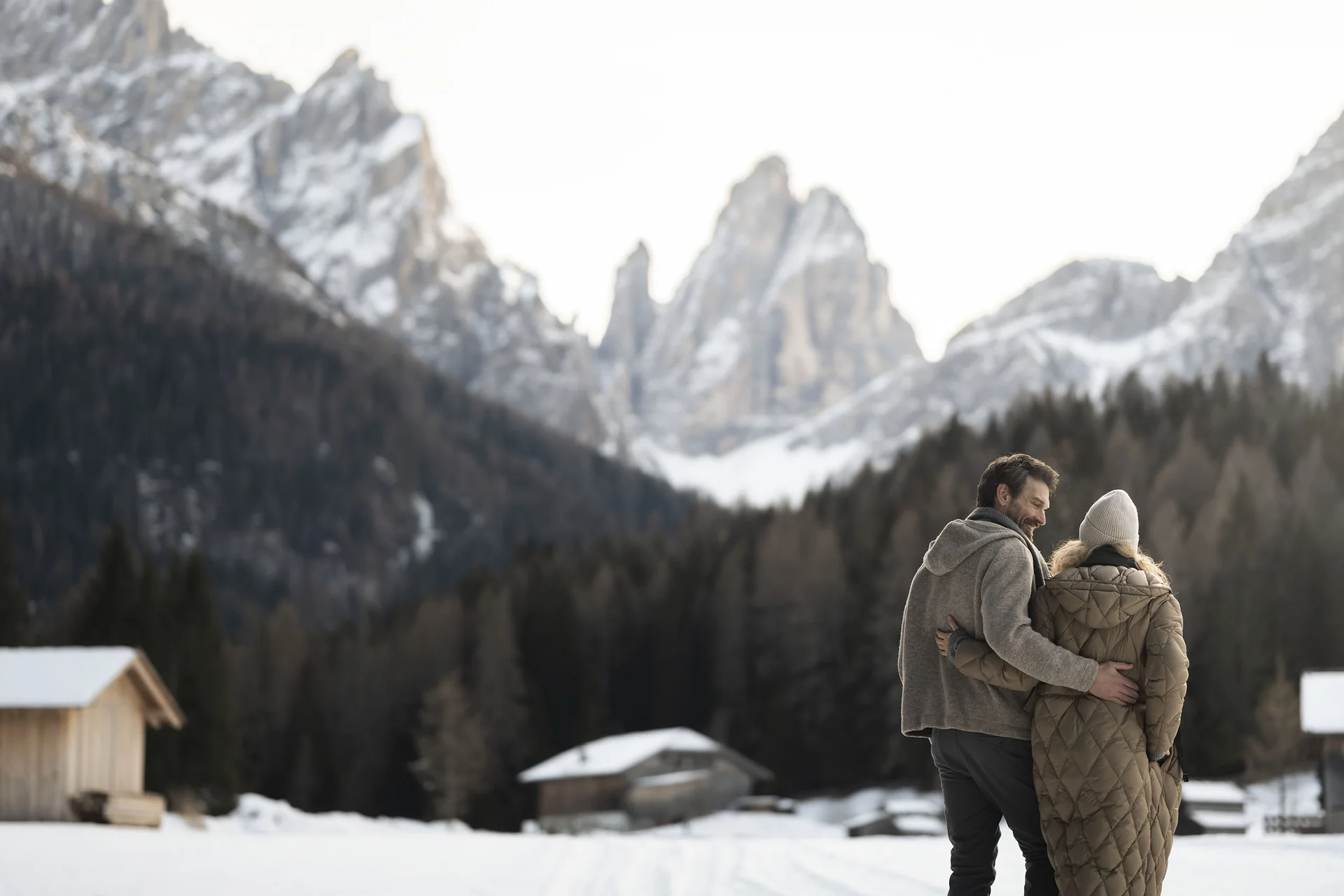 Hotel Monika: eine neue Dimension von Luxus Paar umarmt sich in verschneiter Berglandschaft mit Holzhütten