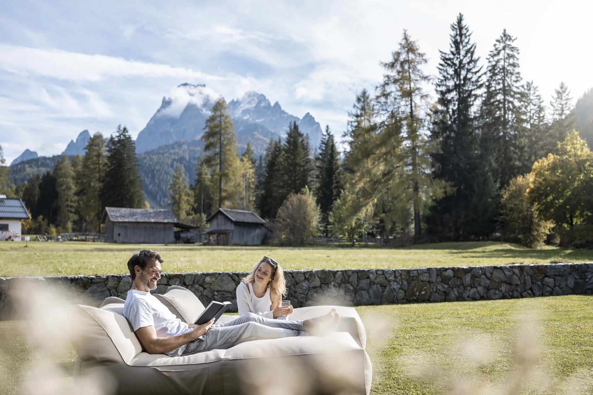 Hotel Monika in Sexten: Wohlbefinden inmitten der Dolomiten Paar entspannt auf Couch im Garten mit Bergkulisse und Häusern im Hintergrund