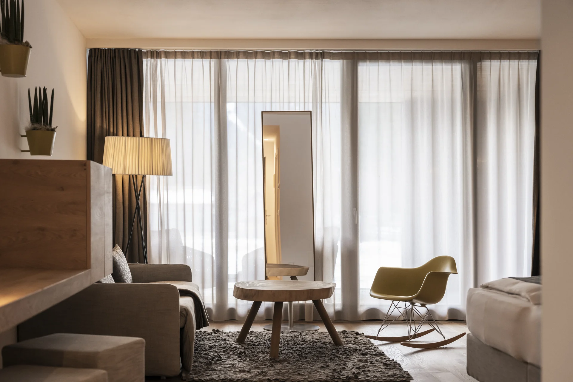Hotel Monika in Sexten: Wohlbefinden inmitten der Dolomiten Gemütliches Wohnzimmer mit Schaukelstuhl, Sofatisch und großem Spiegel vor Gardinen