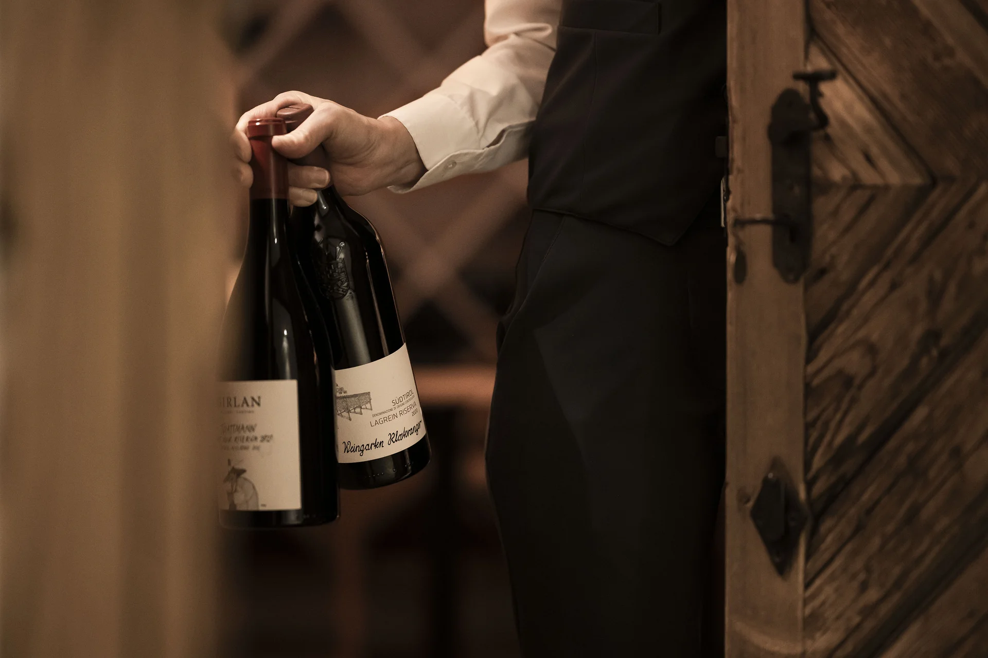 Hotel Monika in Sexten: Wohlbefinden inmitten der Dolomiten Mann trägt zwei Flaschen Wein in einem Weinkeller