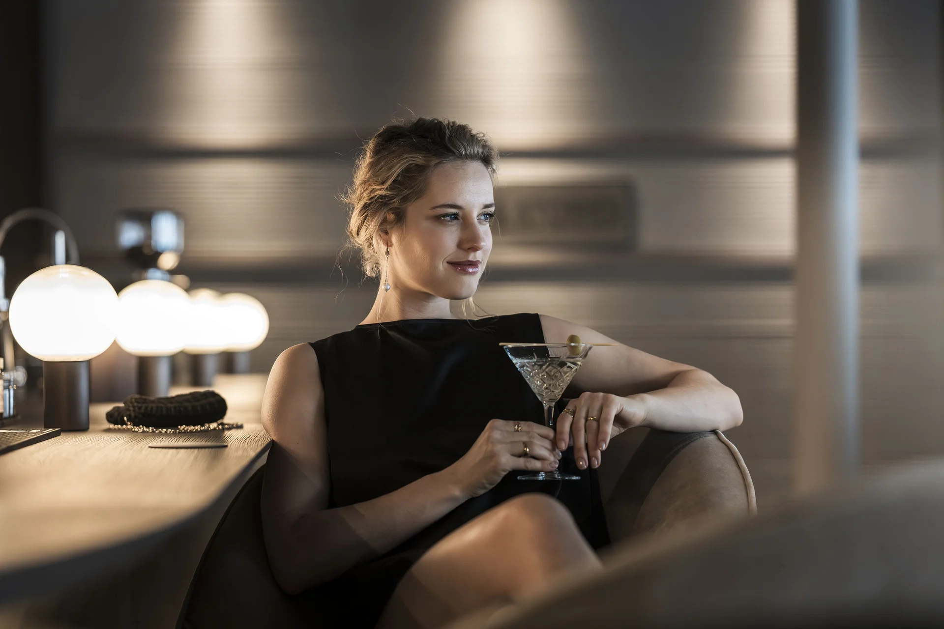 Hotel Monika in Sexten: Wohlbefinden inmitten der Dolomiten Frau in schwarzem Kleid sitzt an Bar mit Cocktailglas und Oliven