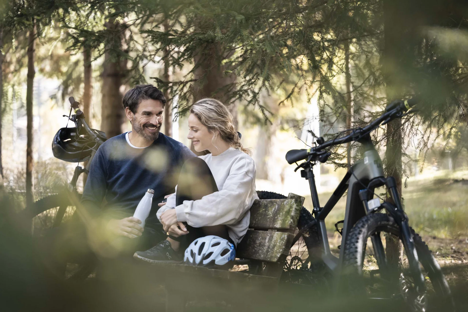 Sommer in den Bergen. In Ihrem Wanderhotel in den Dolomiten. Paar macht Pause auf einer Bank im Wald mit Fahrrädern im Hintergrund