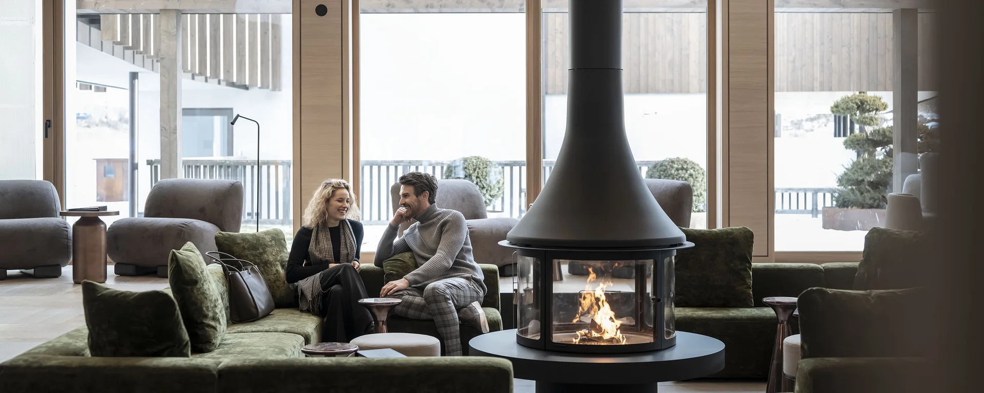 Hotel Monika in Sexten: Wohlbefinden inmitten der Dolomiten Paar sitzt auf grünem Sofa vor modernem Holzofen in hellem Wohnzimmer