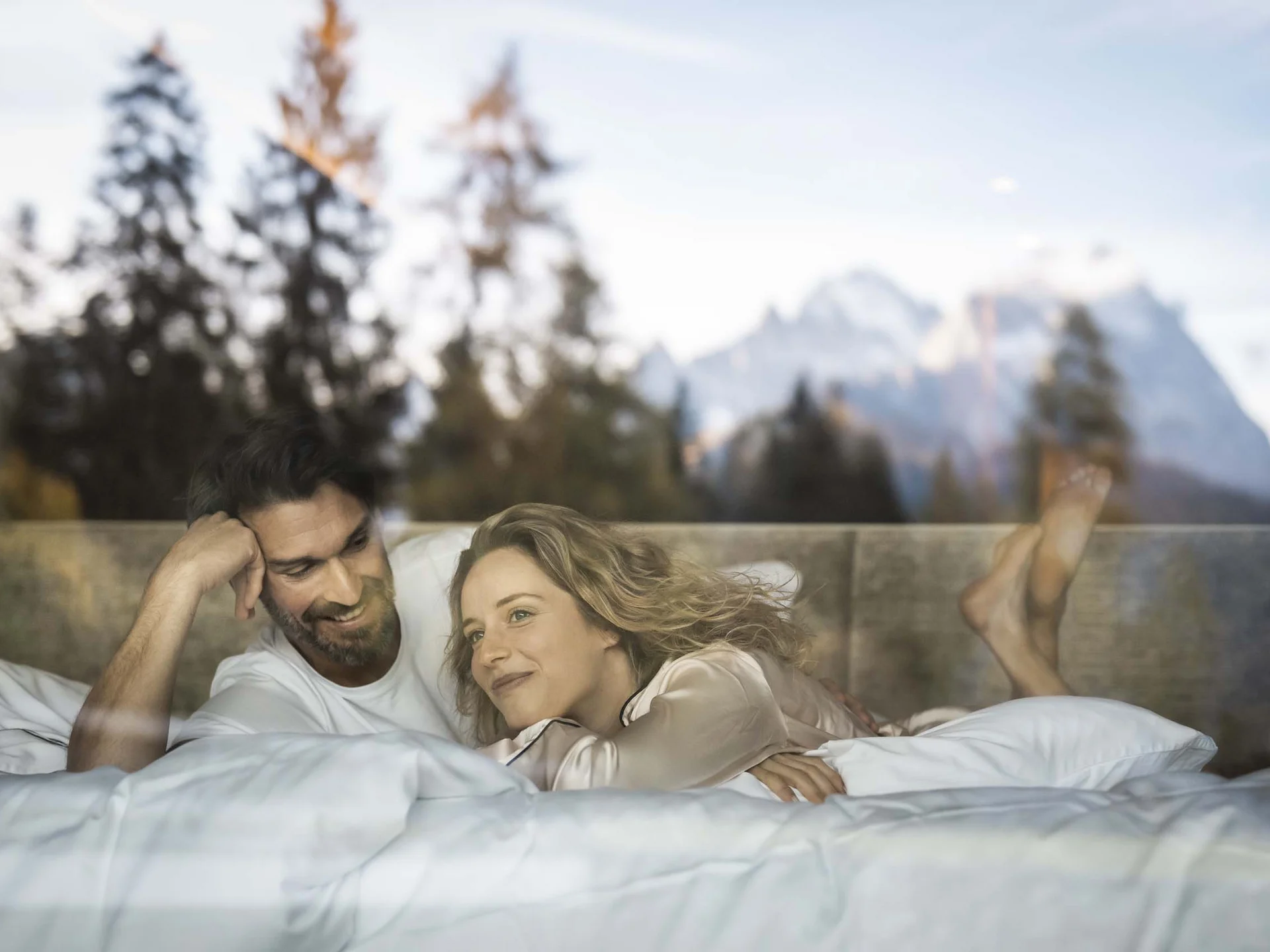Hotel Monika in Sexten: Wohlbefinden inmitten der Dolomiten Paar liegt entspannend im Bett mit Bergblick und Bäumen draußen