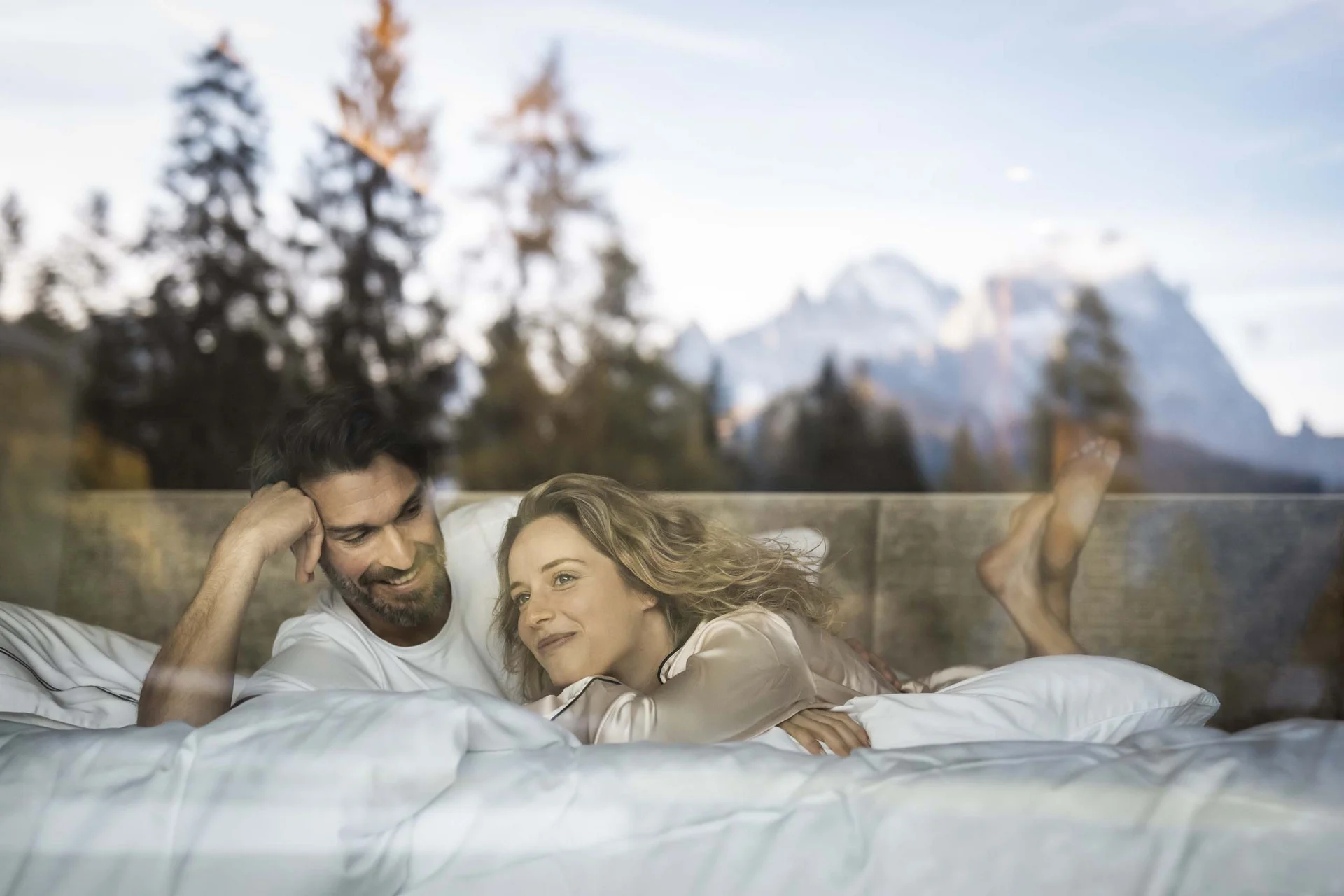 Hotel Monika in Sexten: Wohlbefinden inmitten der Dolomiten Paar liegt entspannend im Bett mit Bergblick und Bäumen draußen