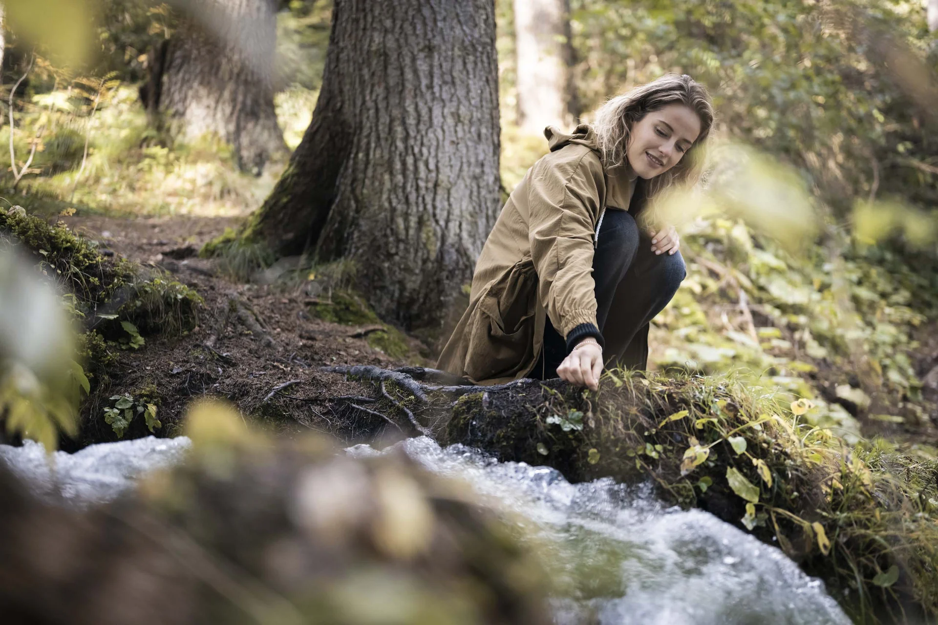 Hotel Monika in Sexten: Wohlbefinden inmitten der Dolomiten Frau sitzt am Waldrand und berührt Wasser eines Baches