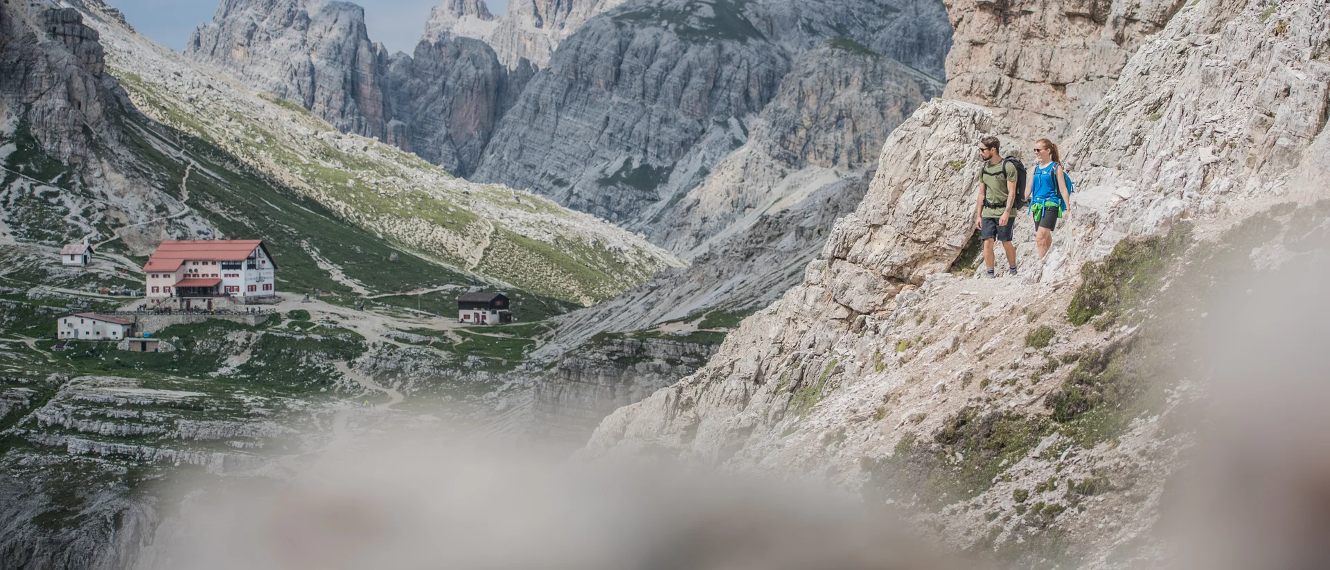 Summer in the mountains. At your hiking hotel in the Dolomites. Two hikers on a rocky mountain trail with mountain huts in the valley